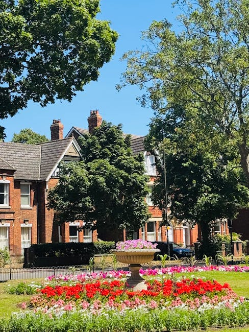 A well-maintained urban garden featuring a colorful flowerbed with red, pink, and white blossoms arranged in neat, blooming clusters. The flowerbed is bordered by a trimmed green lawn and a decorative